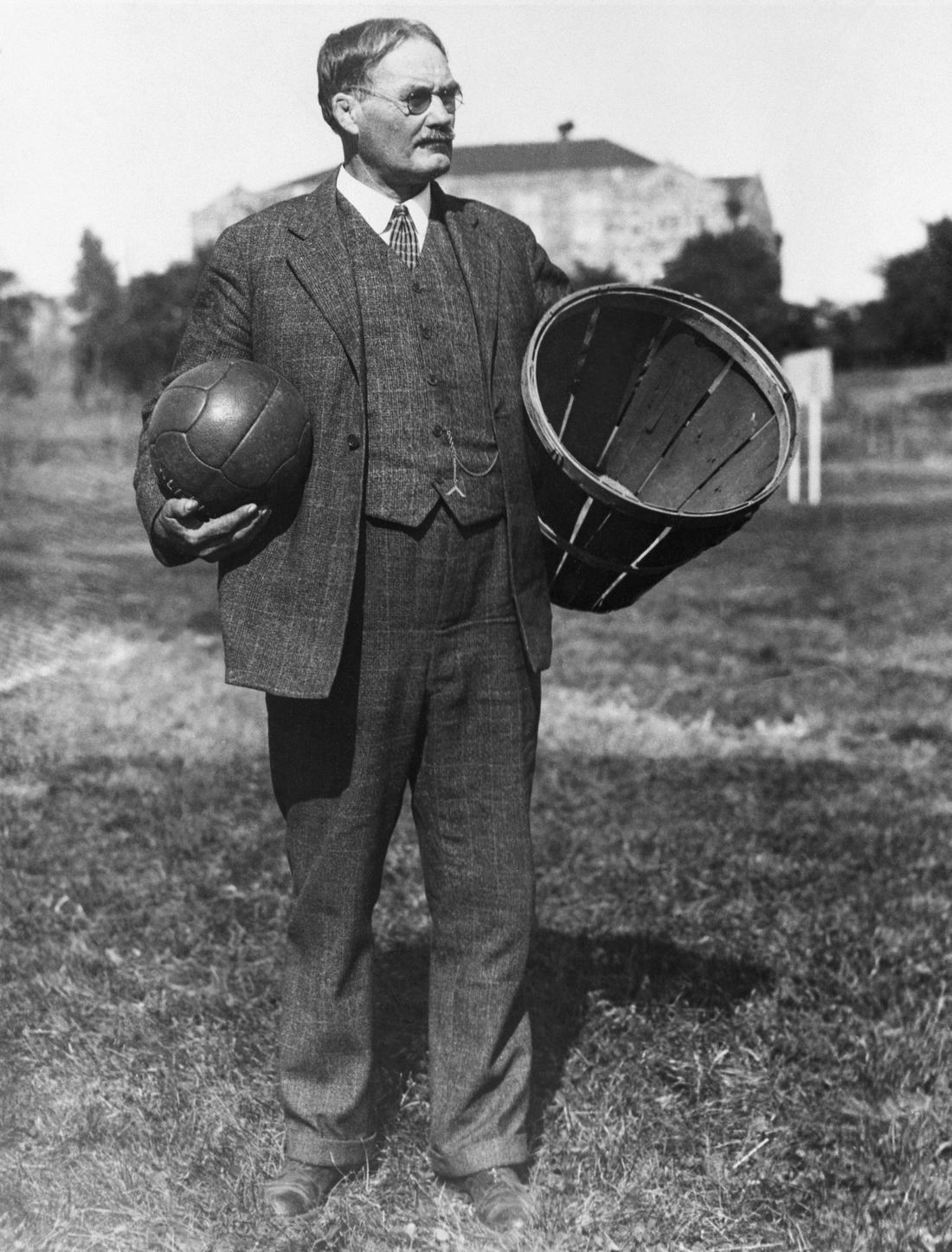 James Naismith standing outdoors holding a ball and an early basket